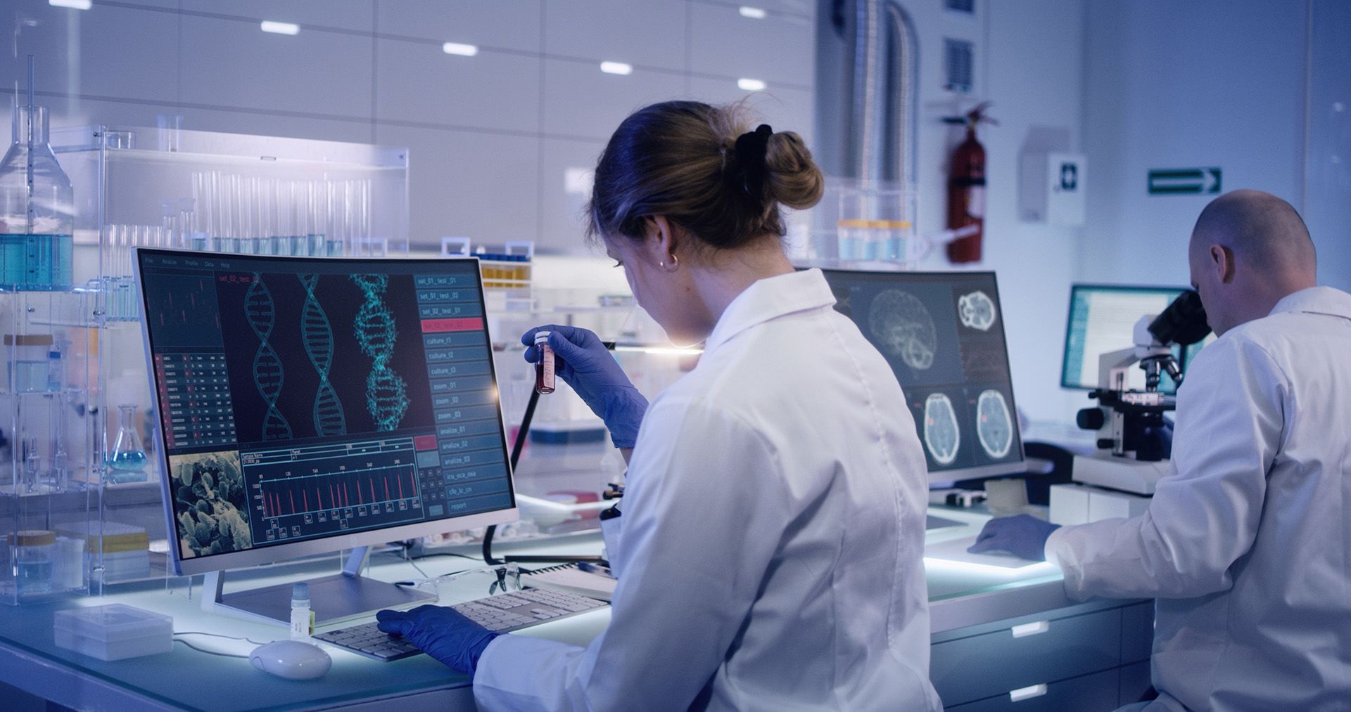 A research team in a modern laboratory. In the foreground a scientist examines DNA on a computer screen while holding a sample in their hand.