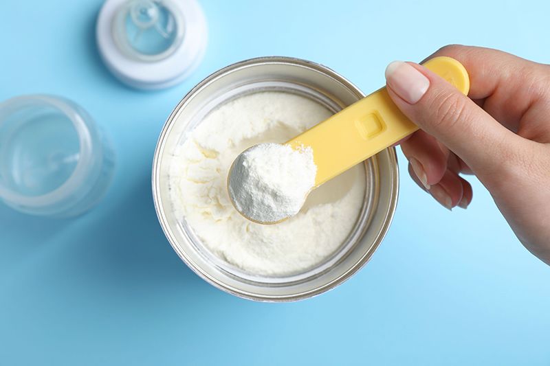 Powdered infant formula being scooped from a can with an infant feeding bottle in the background. 