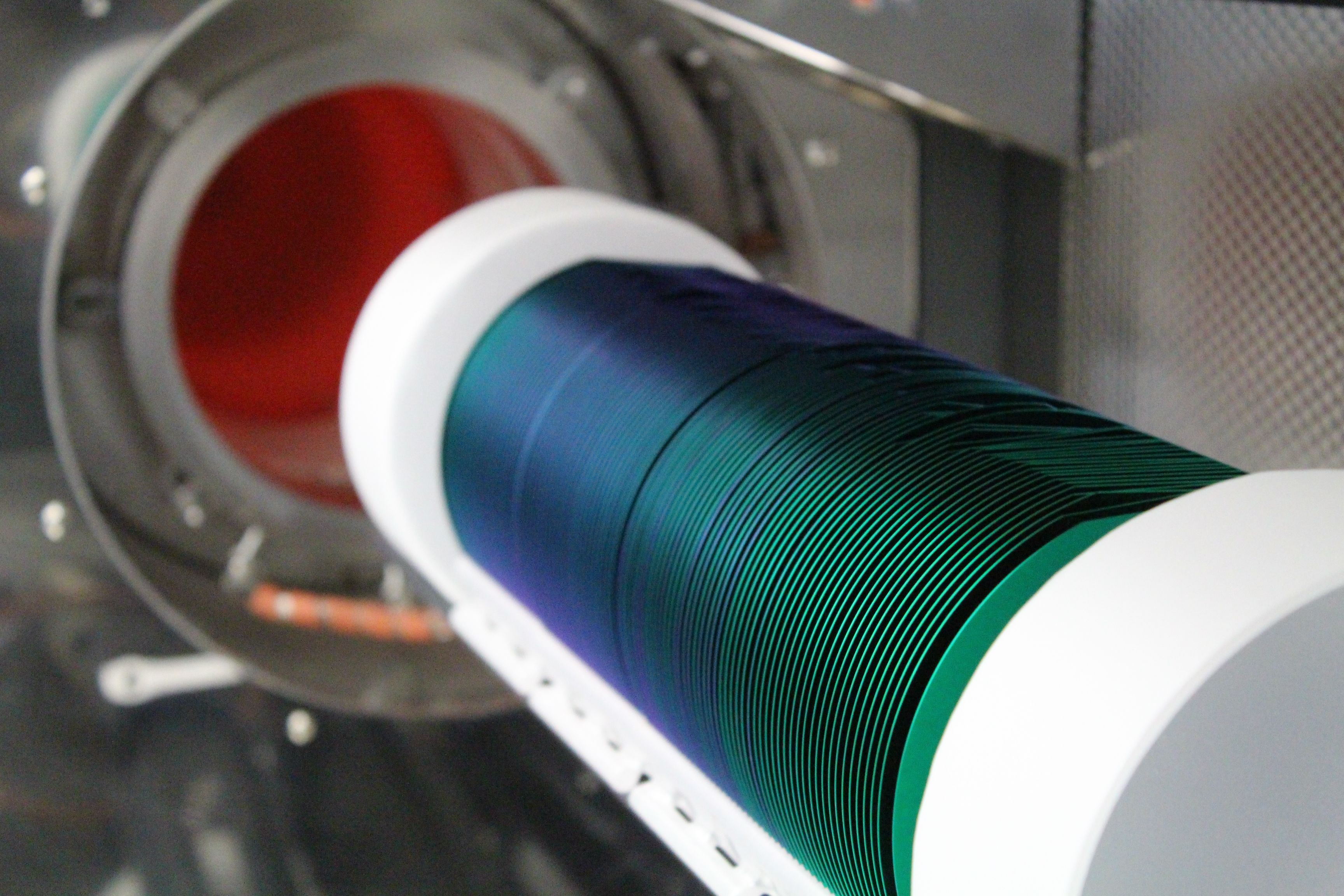 A white horizontal cylinder with the top cut away holds a line of blue green coloured compact discs. The cylinder lies on a diagonal from the front right of the image backwards. In the background a silver tube with a red light is visible.