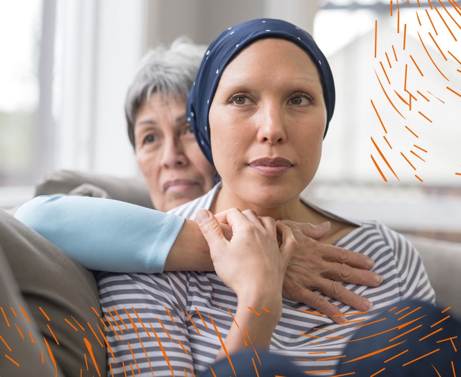 Two female adults sitting on a sofa holding hands. younger is wearing a headscarf due to loss of hair