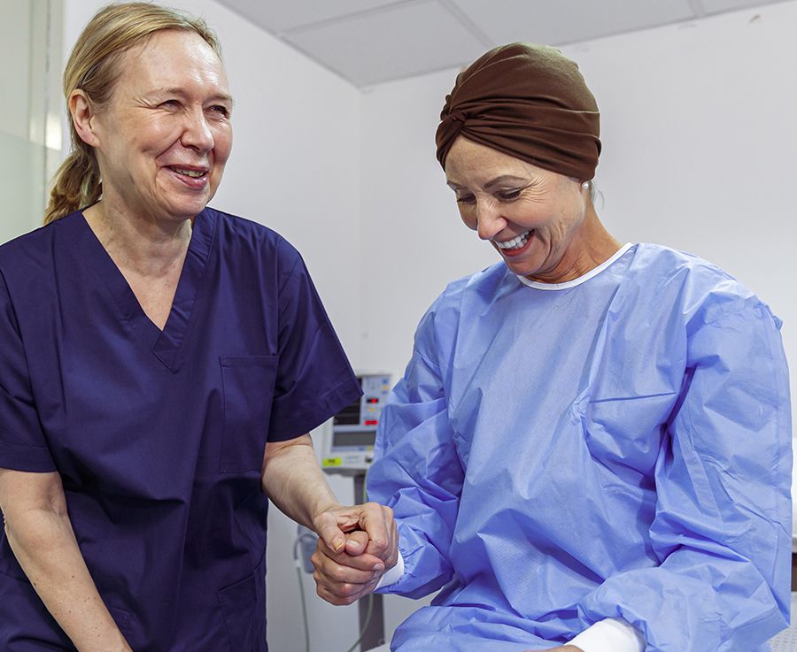 A woman with cancer sits on a hospital bed and interacts with her nurse
