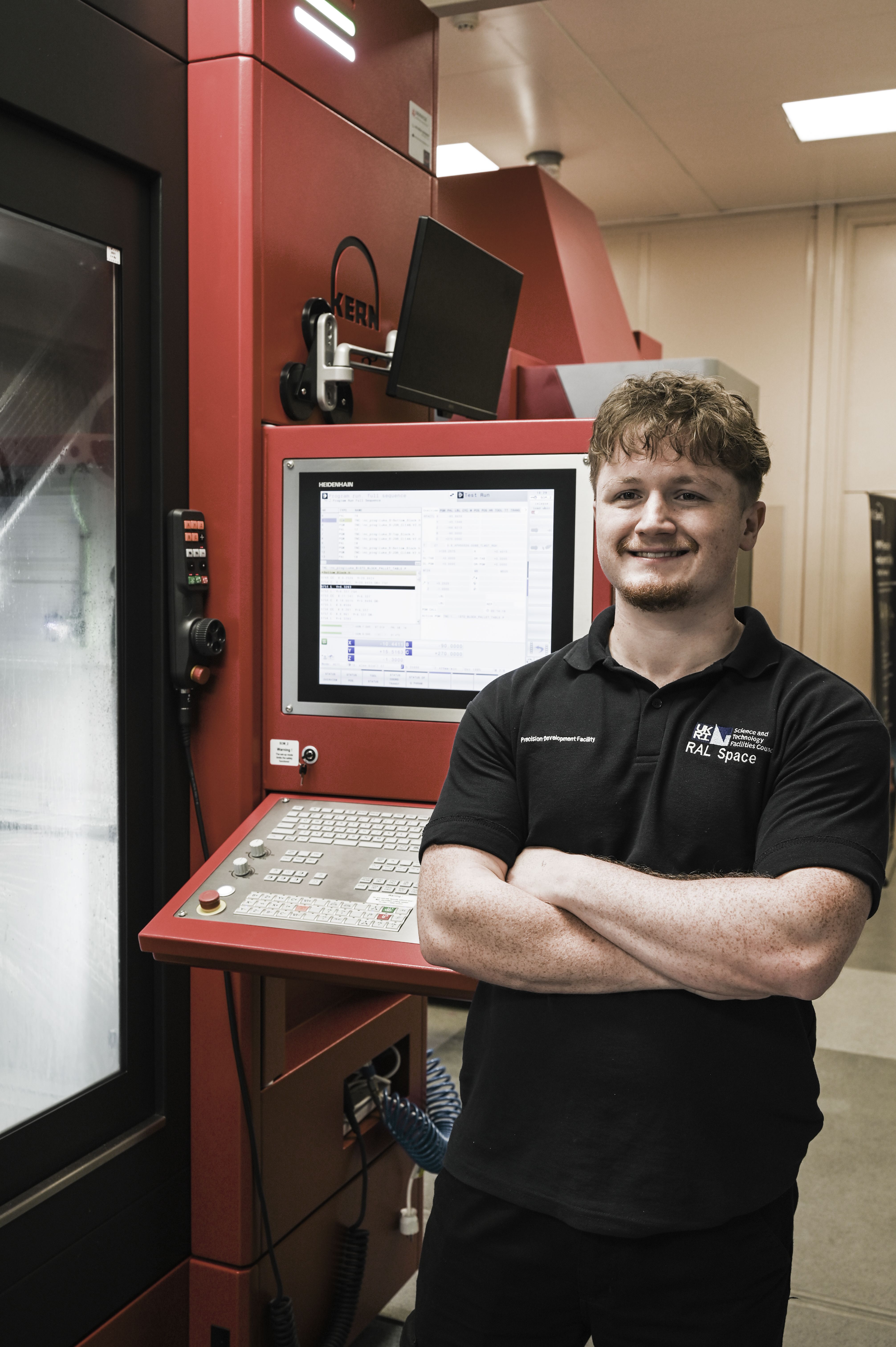Lucas is standing next to some CNC machines and smiling with his arms crossed. He is wearing a RAL Space Precision Development Facility polo shirt.