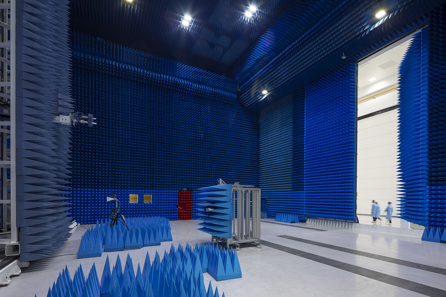 A large room with thousands of blue foam cones lining the walls from floor to ceiling. Some people are seen walking past the open door, but they look very small compared to the vast height of the room.