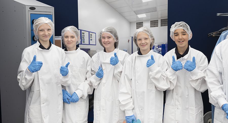 Five school students smiling with their thumbs up during a clean room dress up activity. They are wearing clean room gowns, hairnets and gloves.