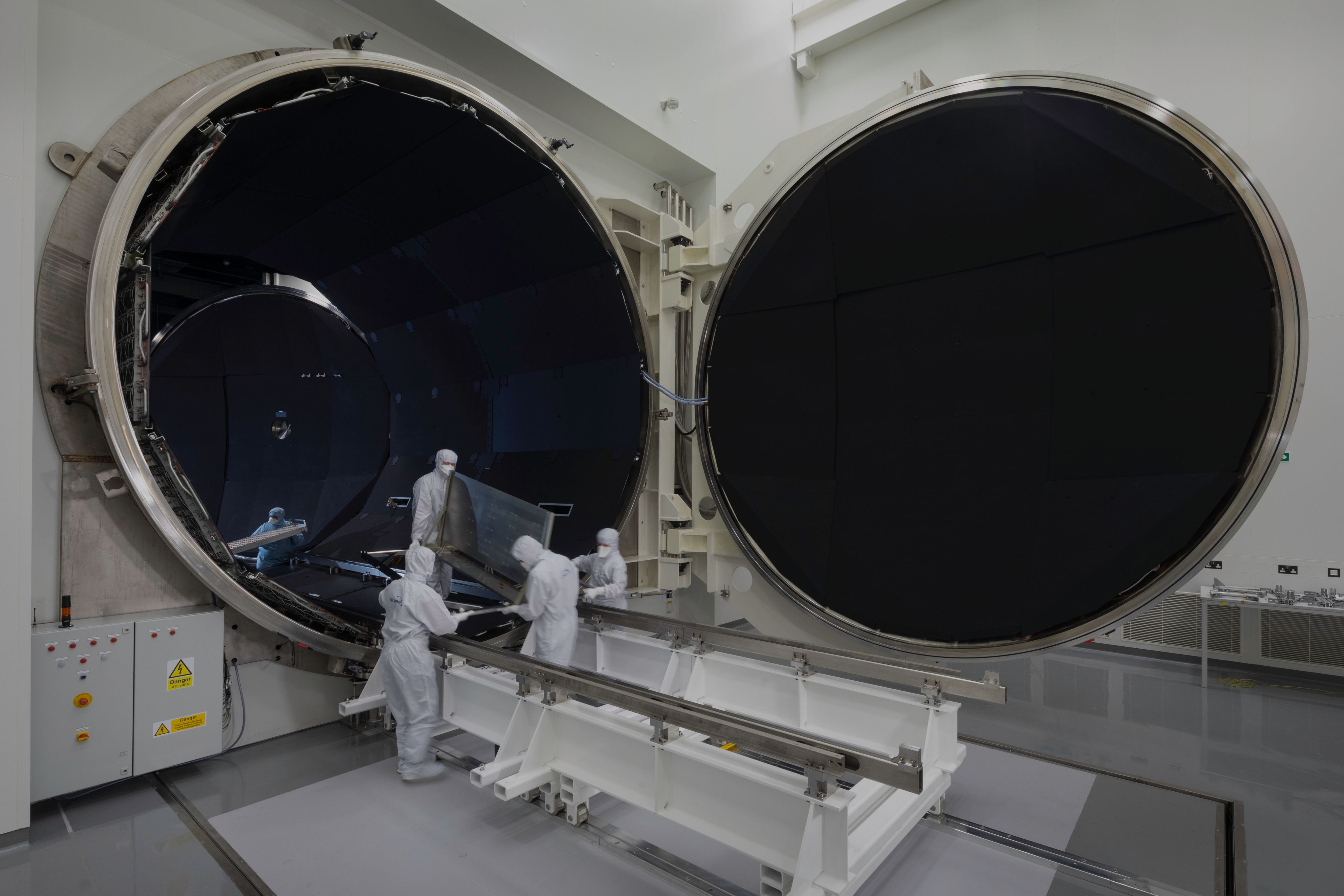 One of RAL Space’s 5 metre diameter thermal vacuum chambers. This is a large cylindrical structure. Four people wearing clean room suits are carrying instrumentation out of the chamber.