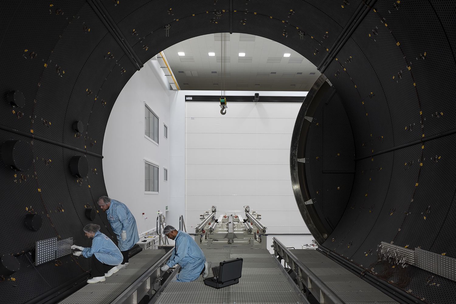 View from inside, looking out of the large space test chamber. The chamber door is circular and 7m wide. Three people wearing clean room clothing are working inside the chamber.