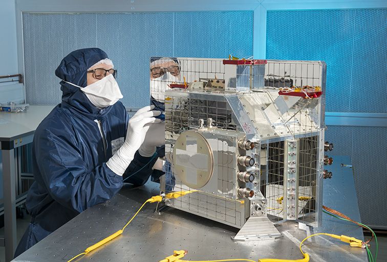 EarthCARE’s BBR – a roughly microwave sized instrument covered in small square mirrors – in a clean room at RAL Space. Someone wearing a clean room suit, mask and gloves is inspecting a small part of the instrument.