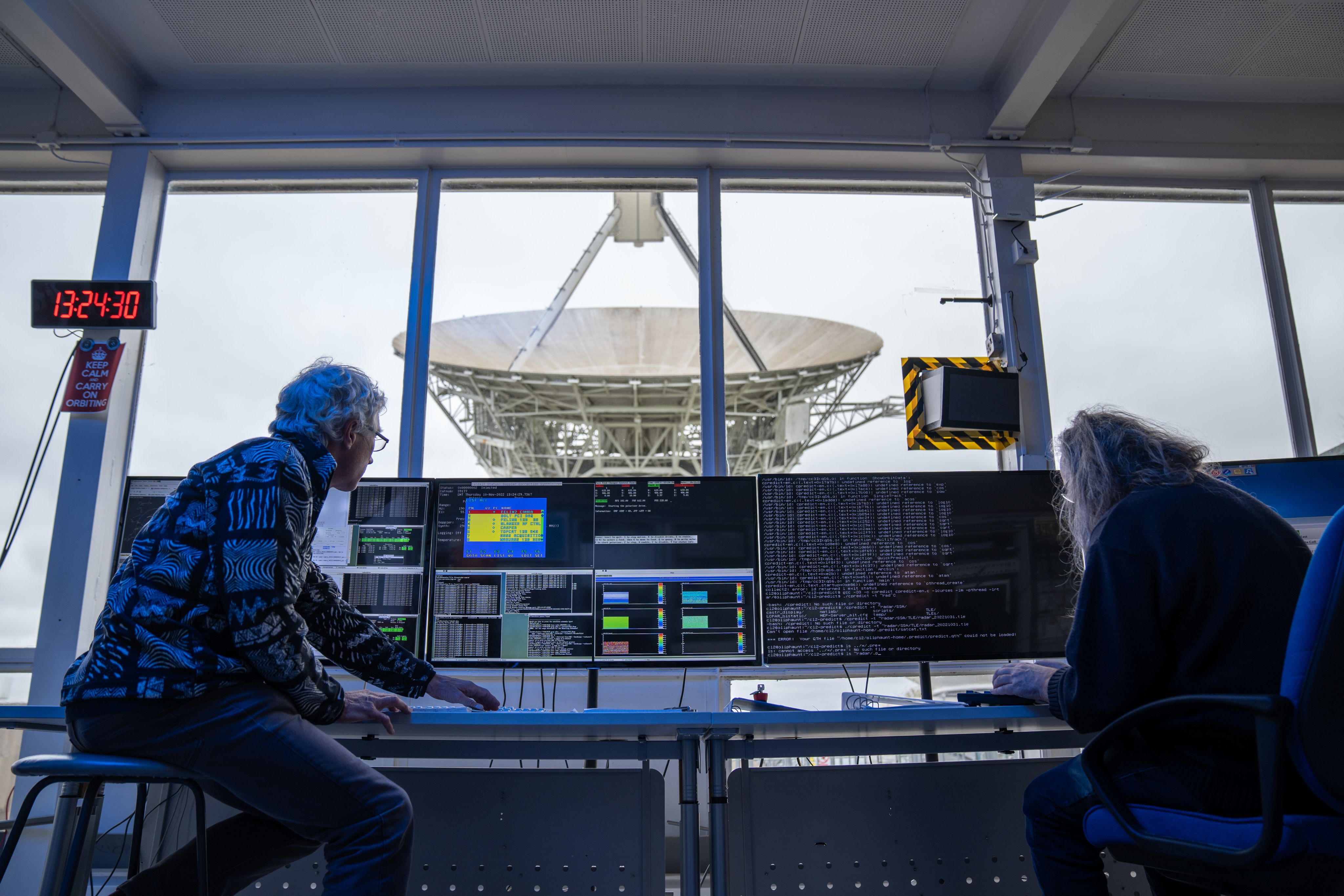 View from inside the Chilbolton Observatory control room. There is one long horizontal window, out of which you can see a large radar dish. A man and woman are sitting with their backs to the camera, at a desk with four PC screens showing various data and code for the radar.