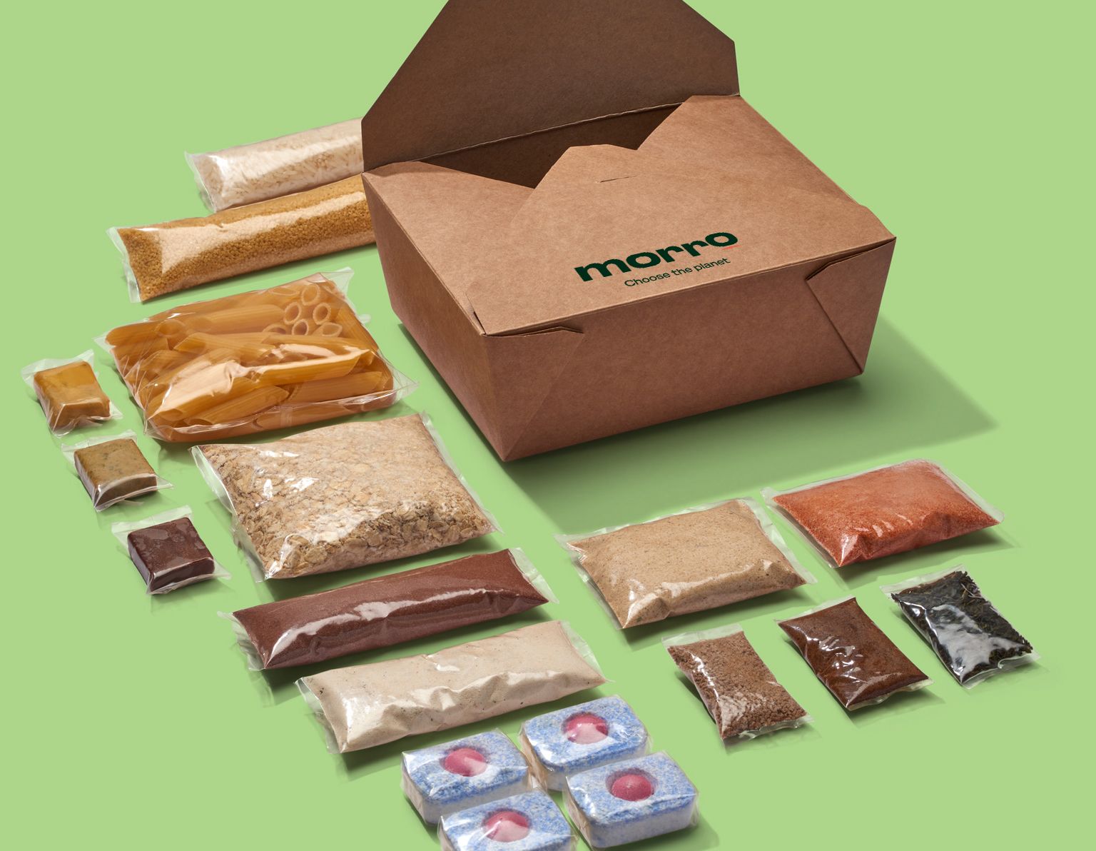 A sample of products from Xampla’s consumer brand Morrow. This consists of individually wrapped packets of dried foods, pasta, oats, grains, powdered spices, stock cubes along with sauces, and dishwasher tablets. Each sample is completely sealed in clear packing at the specific size to hold the volume of the product, to reduce waste. On the bright green surface these samples are spread-out on, there is also a small brown Morrow branded box, to pack them into.