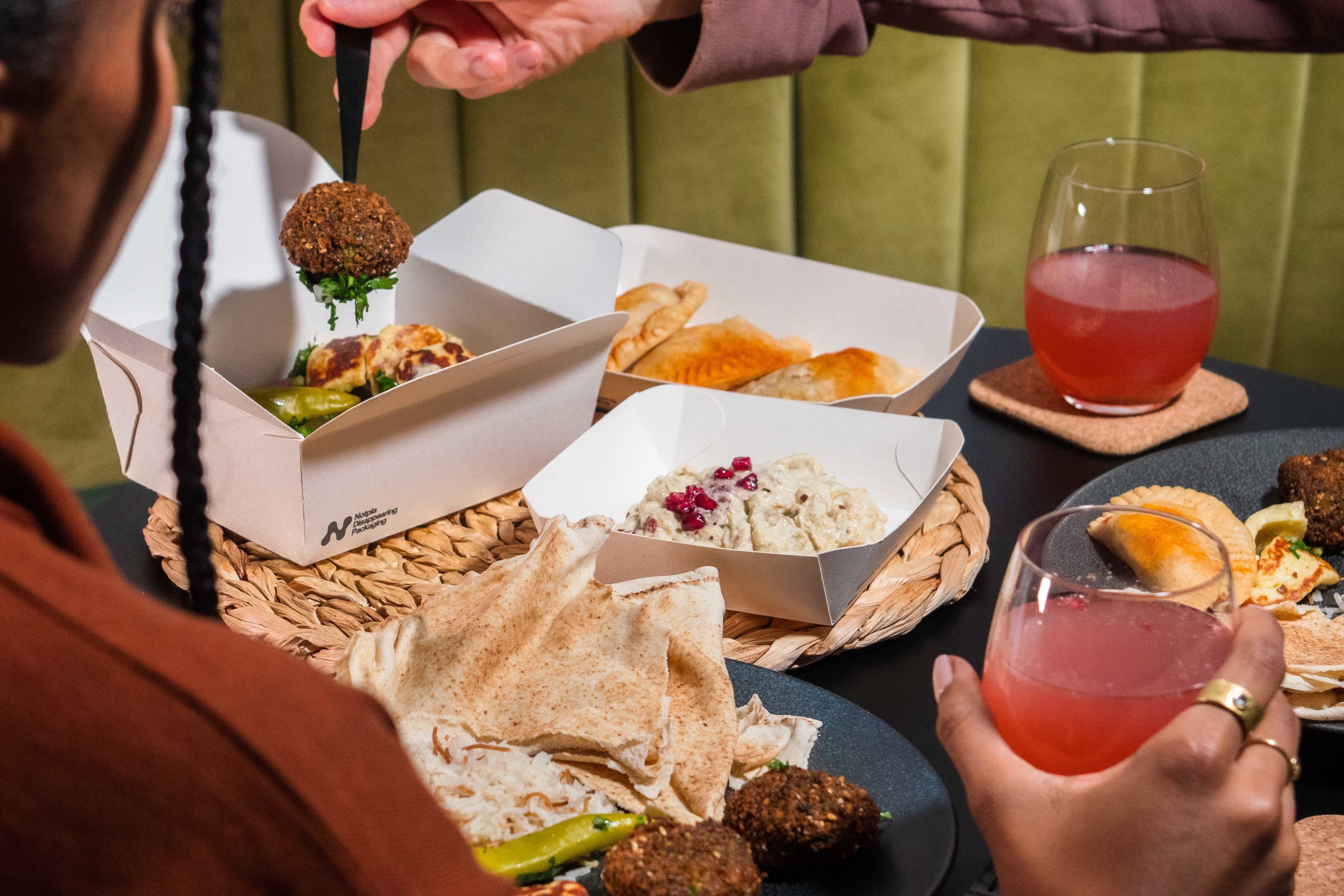 Close up image of a couple at home sitting around a small dining table enjoying drinks and a takeaway meal. The meal is packaged in three white disposable food containers, a box and two open trays, which have been developed by Notpla.