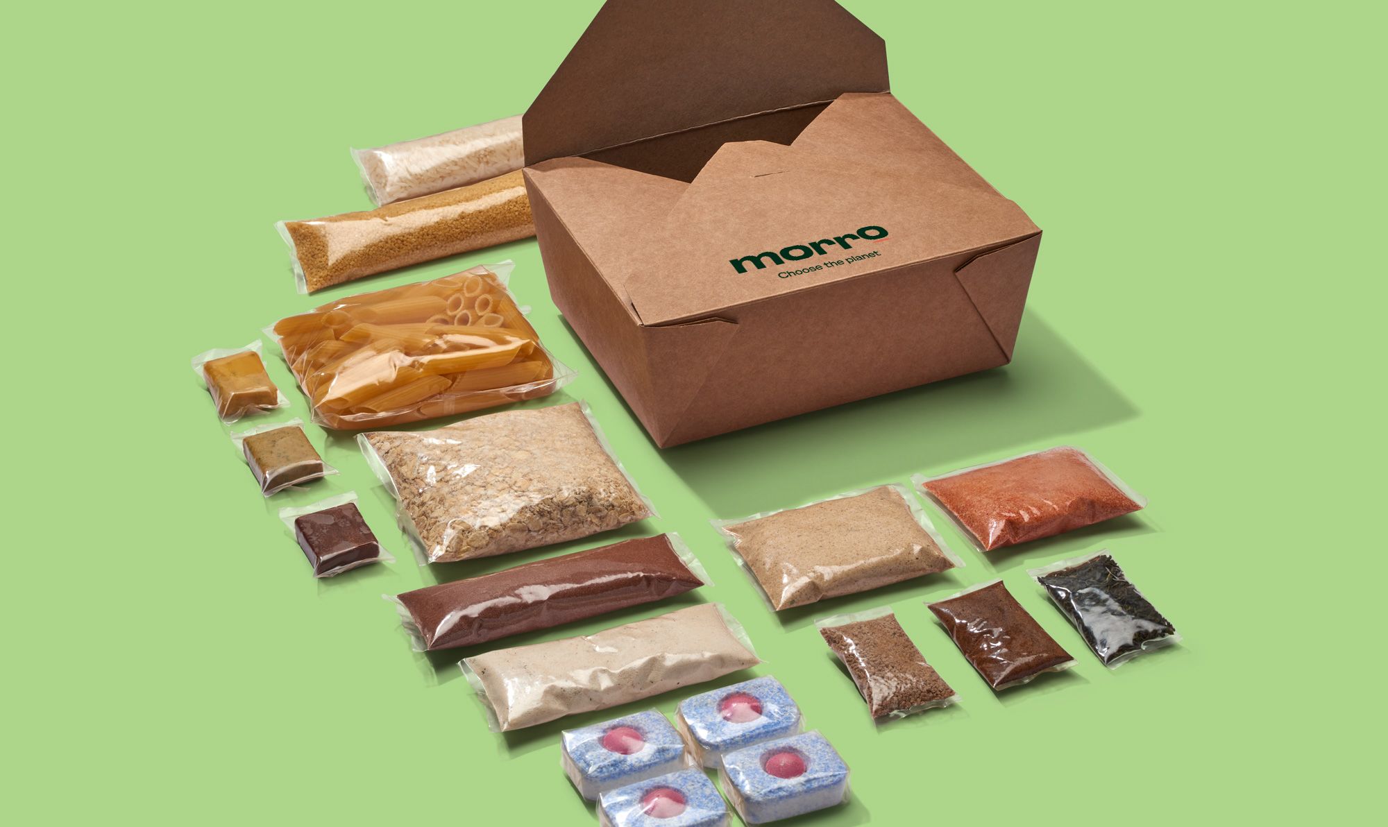 A sample of products from Xampla’s consumer brand Morrow. This consists of individually wrapped packets of dried foods, pasta, oats, grains, powdered spices, stock cubes along with sauces, and dishwasher tablets. Each sample is completely sealed in clear packing at the specific size to hold the volume of the product, to reduce waste. On the bright green surface these samples are spread-out on, there is also a small brown Morrow branded box, to pack them into. 