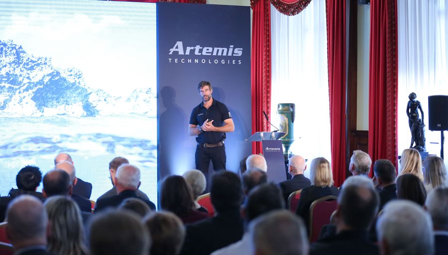 Iain Percy OBE, leader of the project, stands on a stage and addresses an audience with the Artemis logo behind him.