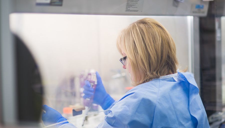 A woman in a paper smock and latex gloves uses a pipette in a laboratory.