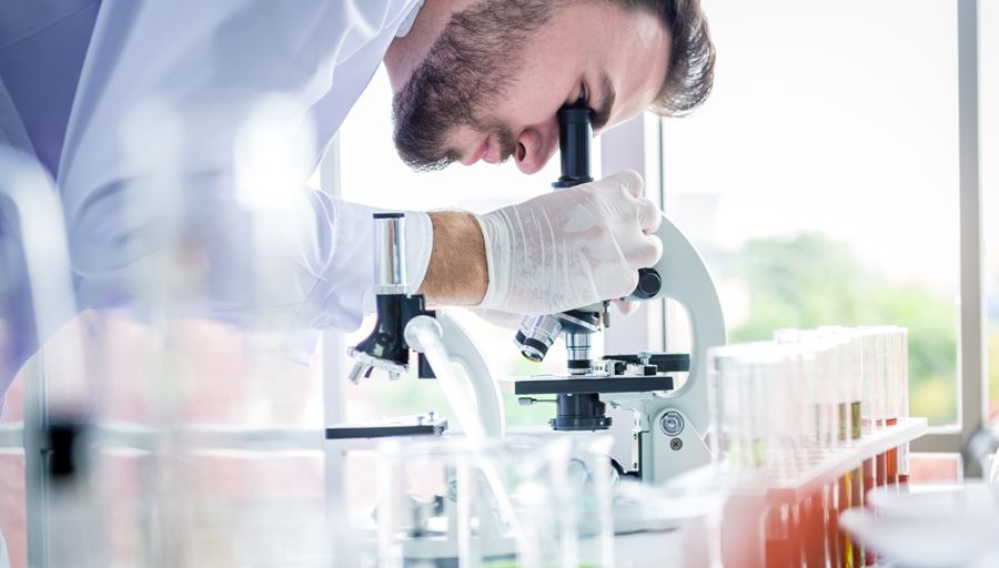 A man in a white coat and latext gloves bends over to look into a microscope.