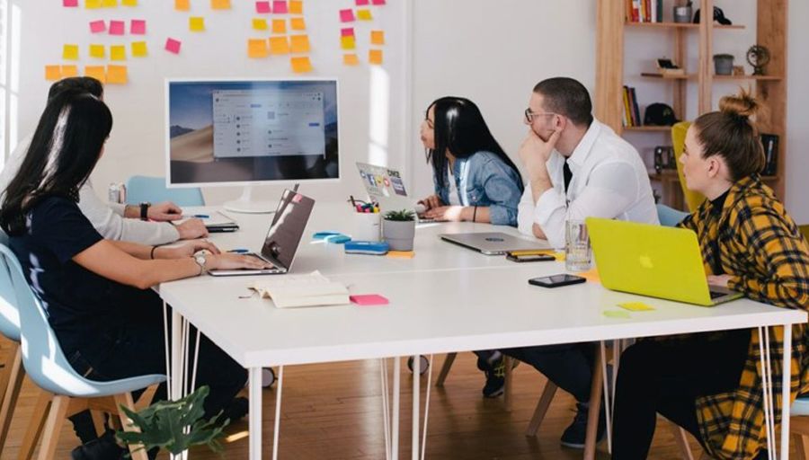Five people sit around a table in a trendy, colourful office and look at a large computer screen. There are many sticky notes on the wall around the screen.
