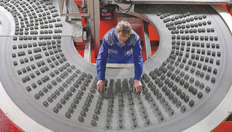 Seen from above, a man in a boiler suit picks up grey blocks from a conveyor belt that curves around him.